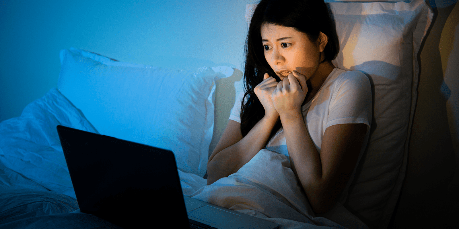 young asian woman watches scary horror film in bed on laptop computer