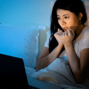 young asian woman watches scary horror film in bed on laptop computer