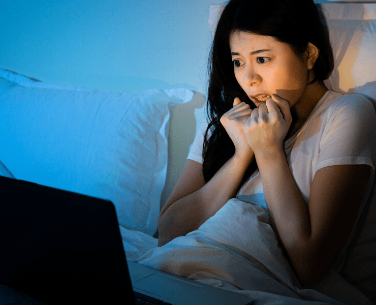 young asian woman watches scary horror film in bed on laptop computer