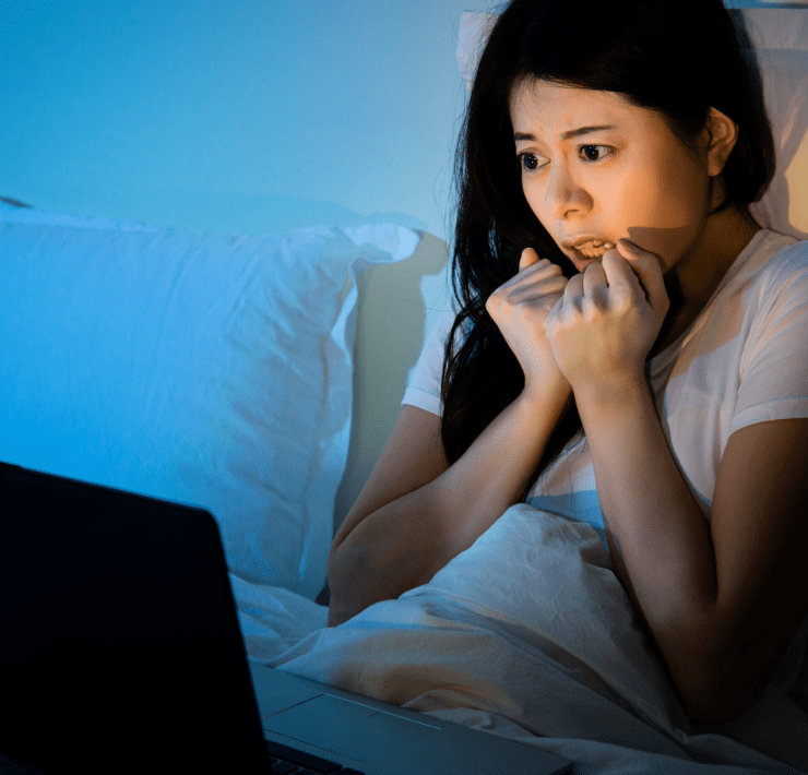 young asian woman watches scary horror film in bed on laptop computer